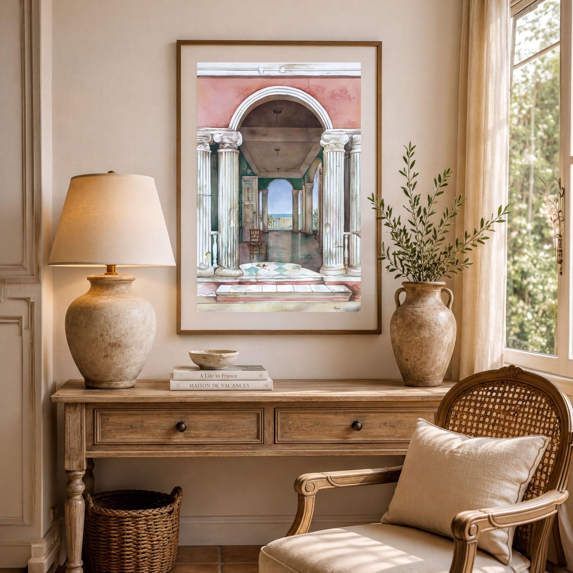 a cozy living room with a wooden table, a lamp, a woven chair, and a framed painting of an archway. The room has a warm and inviting atmosphere, with natural light coming in through a window and greenery from a plant.