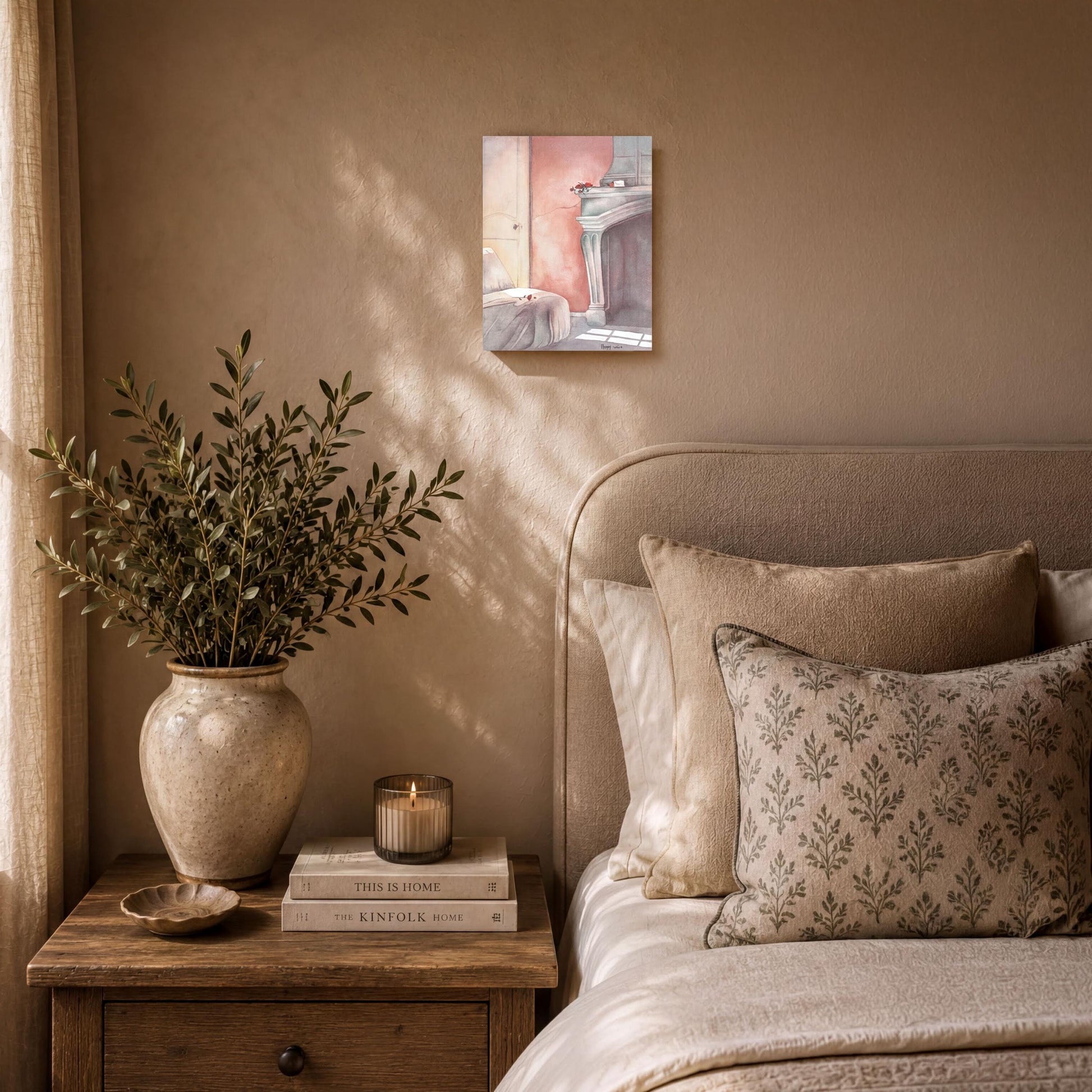 a cozy bedroom scene with a bed, nightstand, and artwork on the wall. The bed is adorned with a white comforter and a patterned pillow, and a vase of greenery sits on the nightstand. A stack of books is also present on the nightstand. The artwork on the wall adds a personal touch to the space.