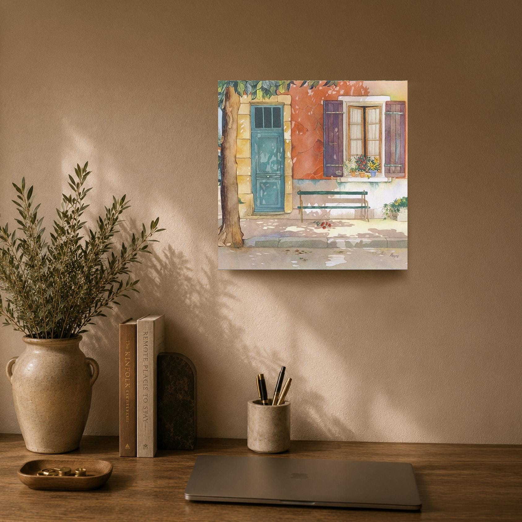 a wooden desk with a laptop, a vase with greenery, and a painting of a house with a blue door and red shutters.